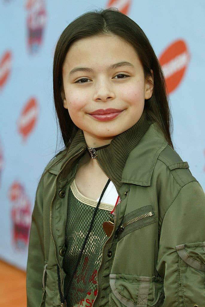 Miranda Cosgrove on the red carpet wearing a green jacket over a mesh top, smiling towards the camera
