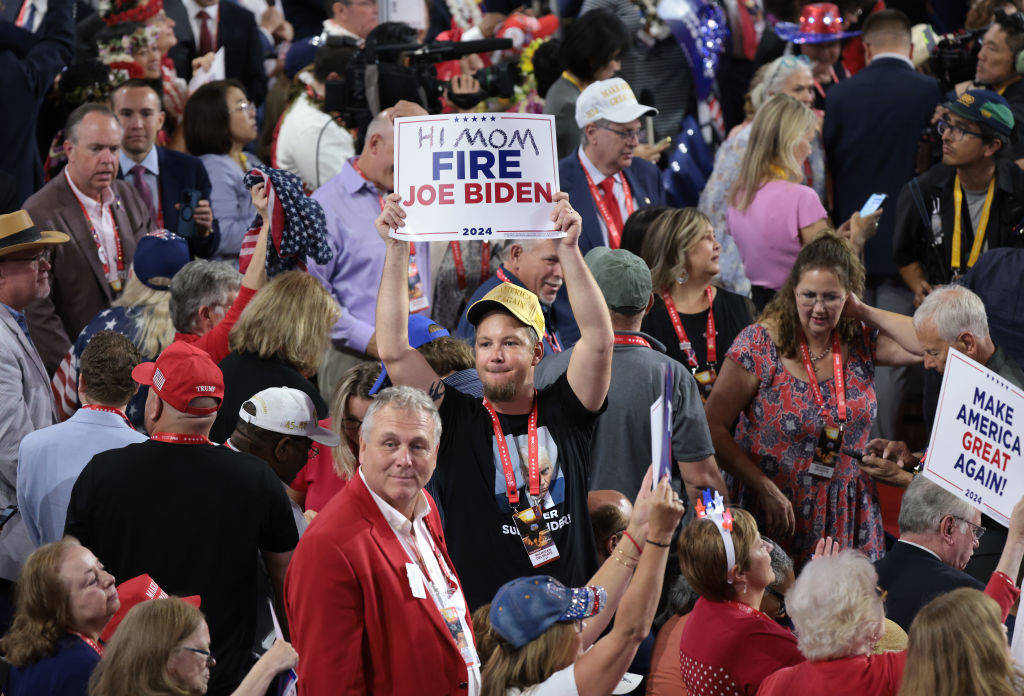 A man in a crowd holds a sign reading &quot;Hi Mom Fire Joe Biden 2024&quot; during a political rally. People around him are wearing various attire with political slogans