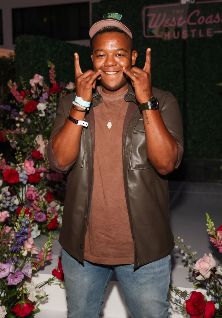 A person smiling, making hand gestures, wearing jeans, a brown shirt, a brown jacket, a watch, and a green cap, standing in front of a floral display and West Coast Hustle sign