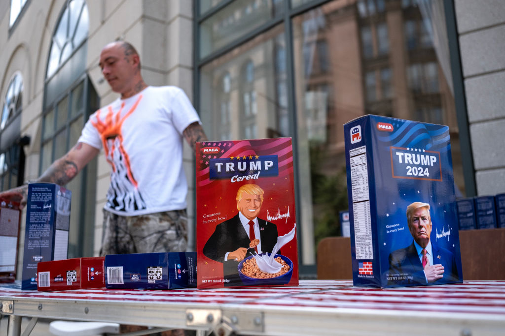A person stands behind a table displaying various Trump-themed cereal boxes outdoors. The boxes feature illustrations of former President Donald Trump