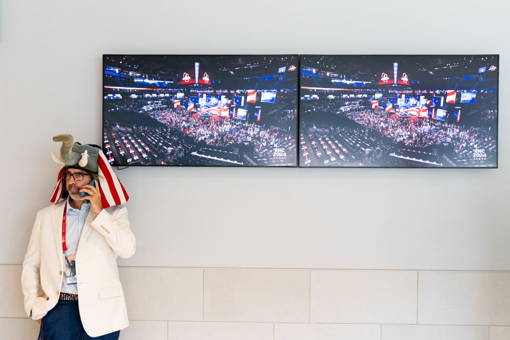 Man wearing a suit, tie, and elephant hat stands in front of two TV screens showing a crowded indoor event