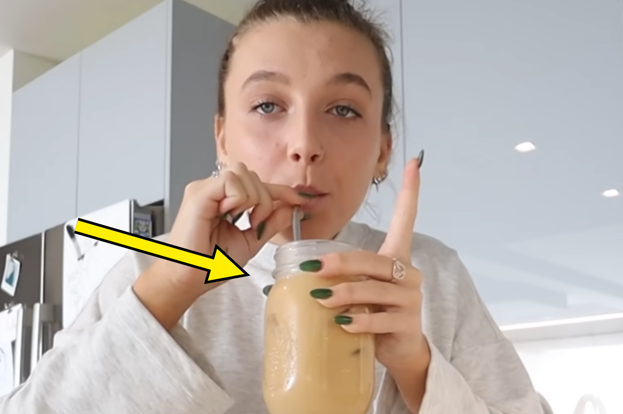 Emma Chamberlain drinks iced coffee from a mason jar straw while raising one finger, wearing a cozy, casual outfit