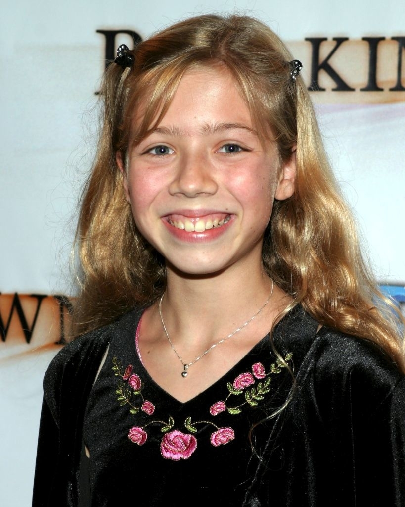 A young girl with long hair, wearing a black dress with floral embroidery, smiles at the camera at an event