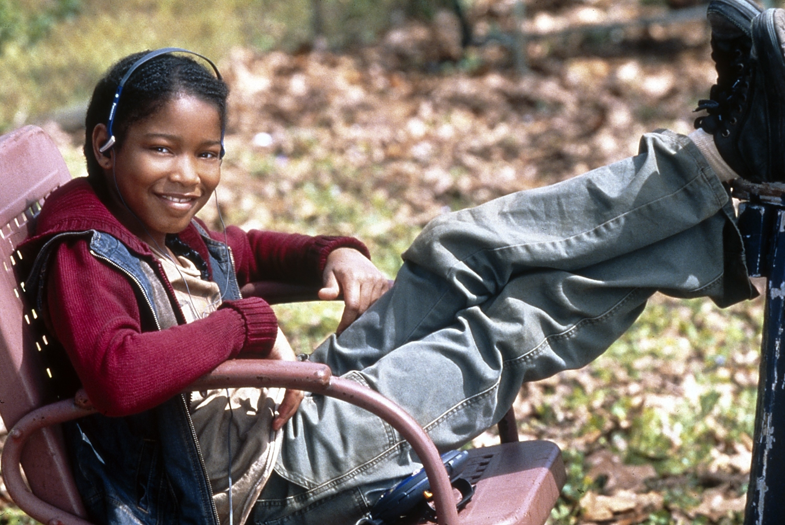 A boy sits relaxed in a chair outdoors with his legs propped up, wearing a sweater over a shirt, pants, and headphones
