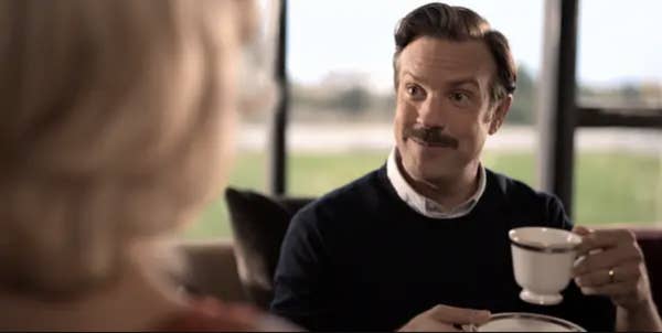 Jason Sudeikis, as Ted Lasso, sits holding a teacup and saucer, smiling and conversing with a blurred person in the foreground