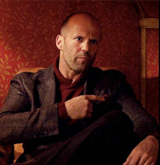 Jason Statham sitting in a chair, wearing a turtleneck sweater and a blazer, gesturing with his hand