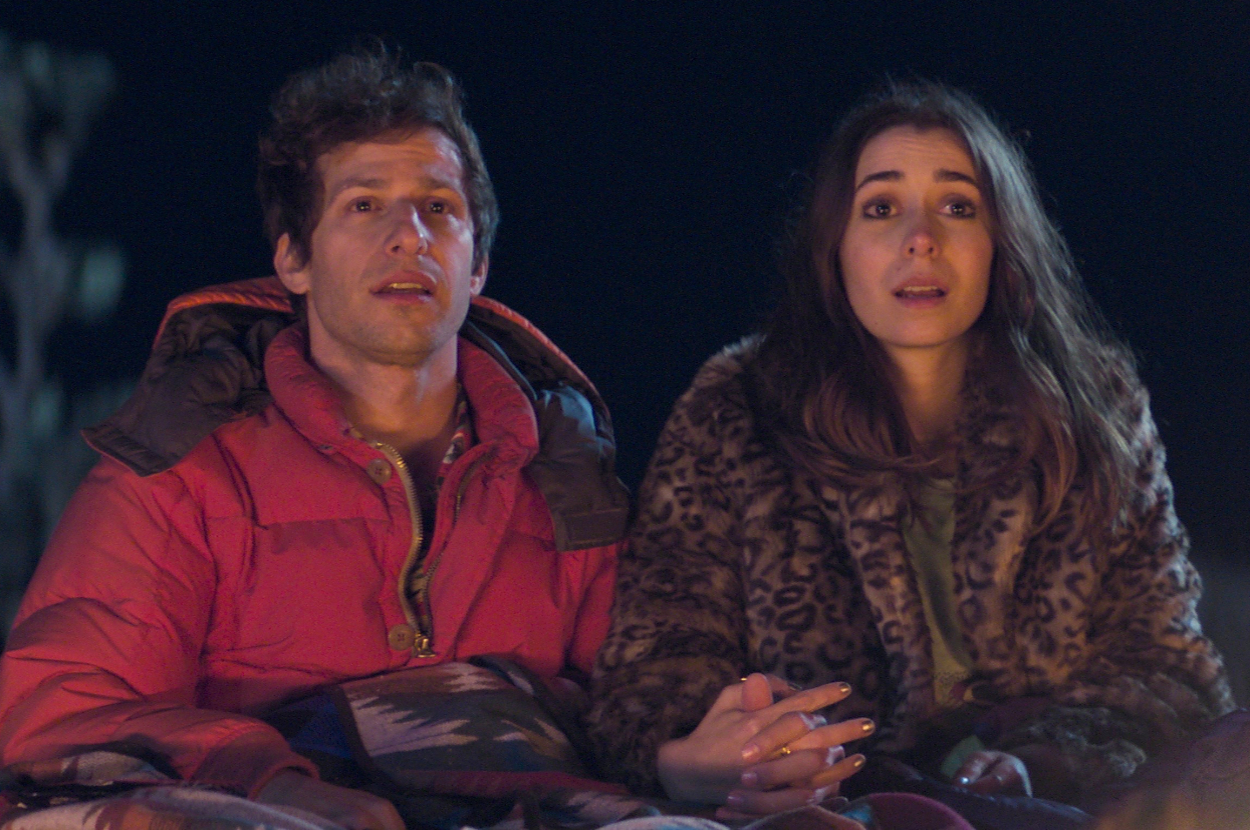 Andy Samberg and Cristin Milioti sitting outdoors, looking intently at something as Nyles and Sarah in Palm Springs