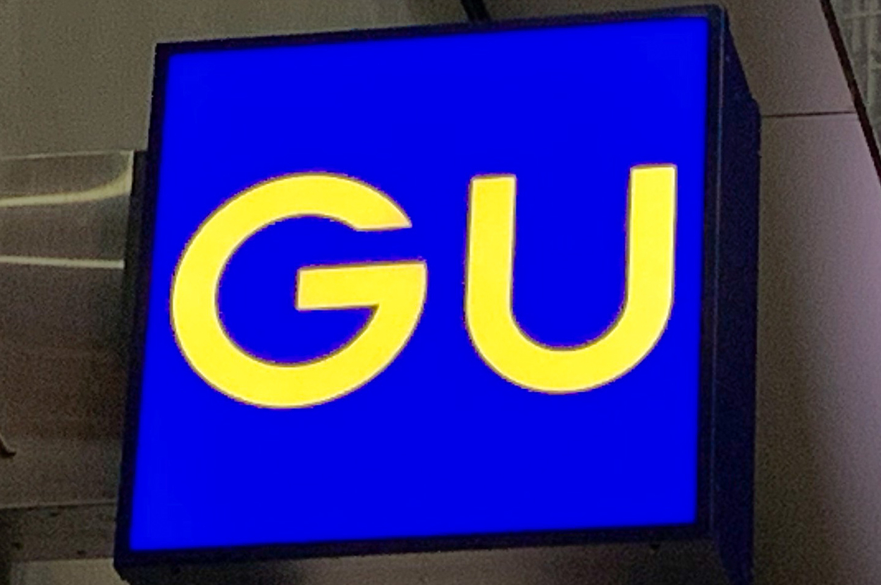 A square blue sign with the yellow letters "GU" is mounted on a building