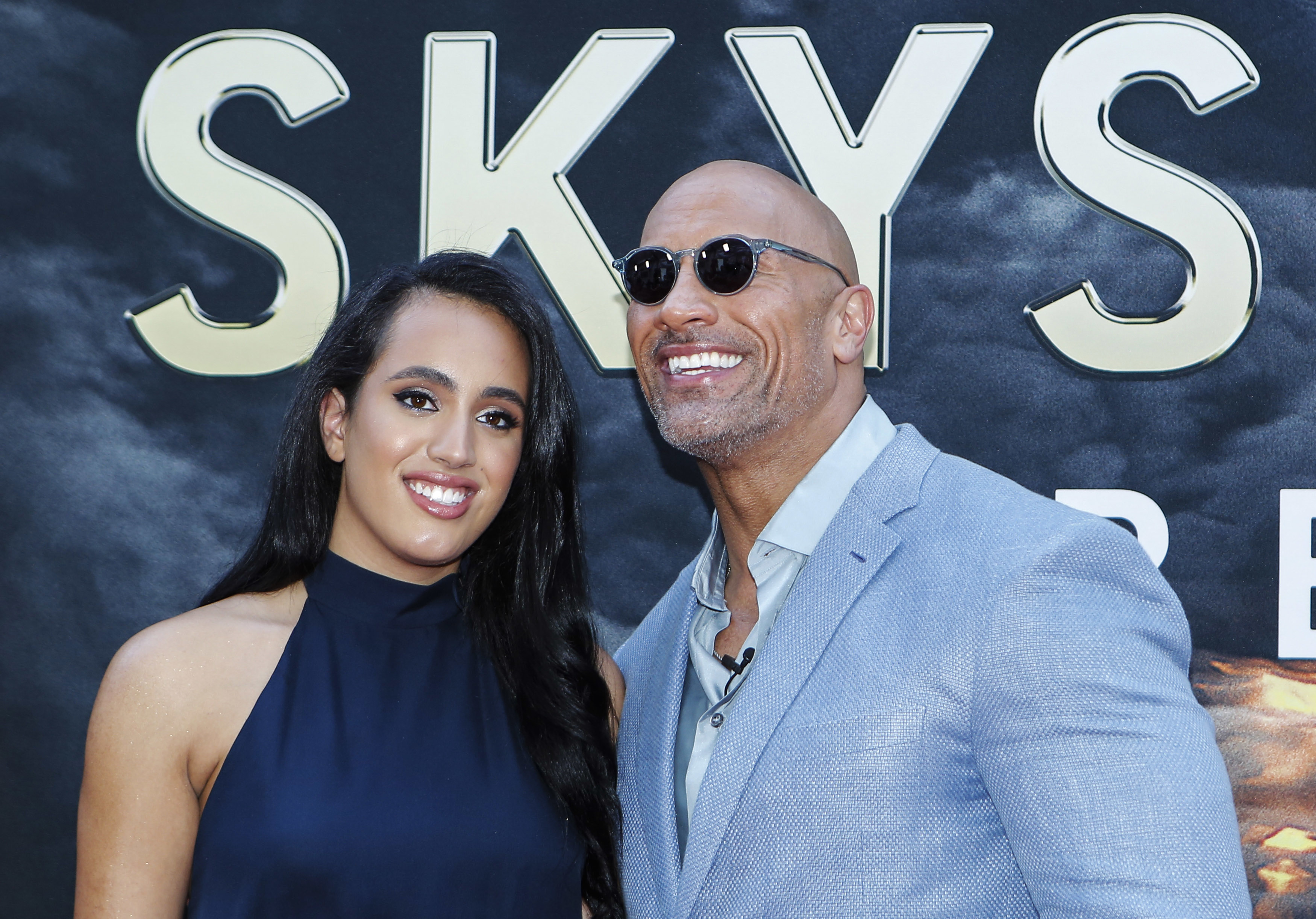 Dwayne Johnson in a light suit and sunglasses poses with his daughter Simone Alexandra Johnson in a dark high-neck dress at a premiere event