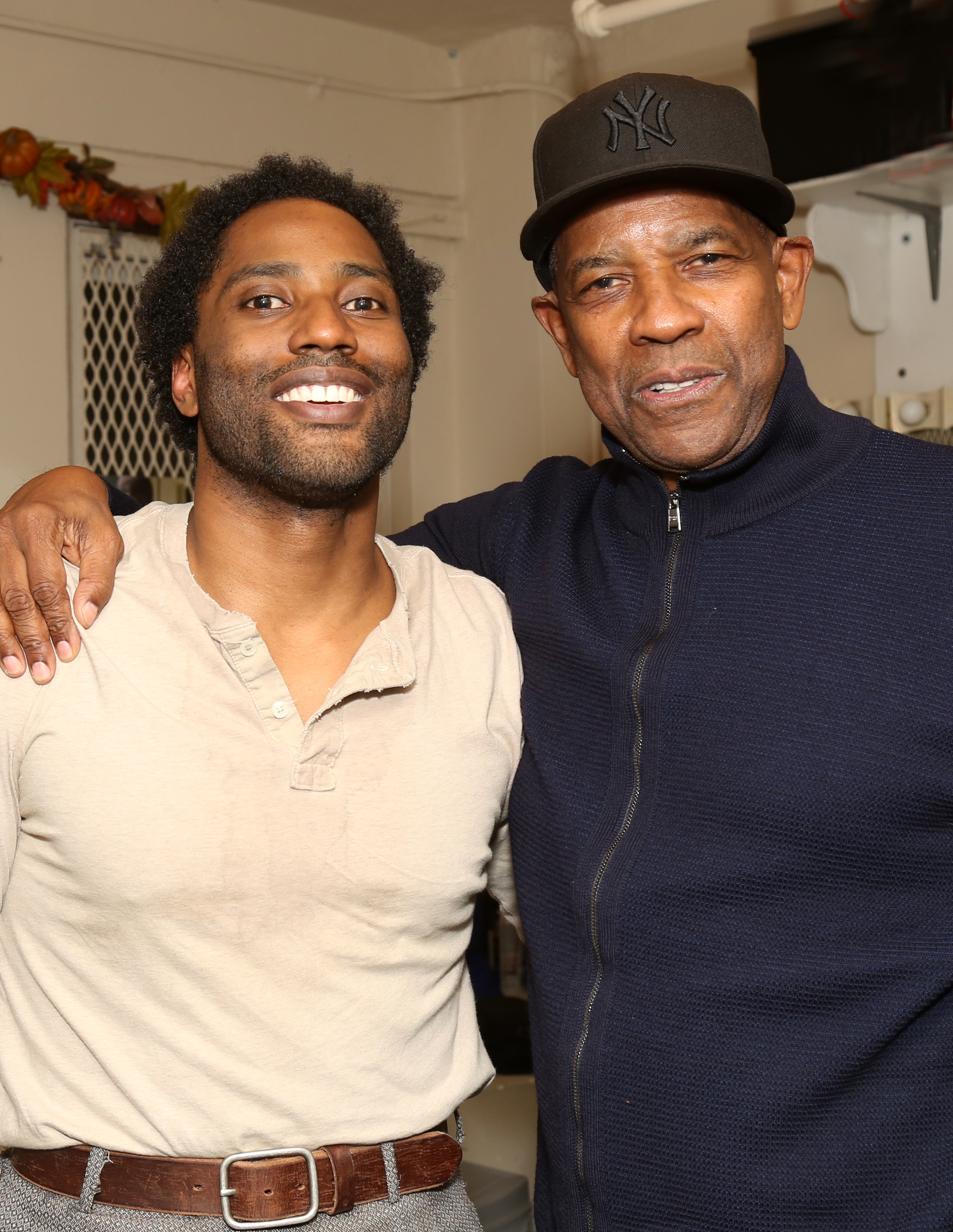John David Washington and Denzel Washington smiling, with Denzel's arm around John David's shoulder. John David is wearing a casual shirt and Denzel a dark jacket and cap