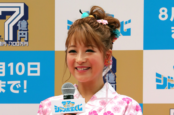 女性が花柄の浴衣を着て、マイクを持って笑顔でカメラに向かっています。彼女の背後にはイベントの広告が掲示されています。