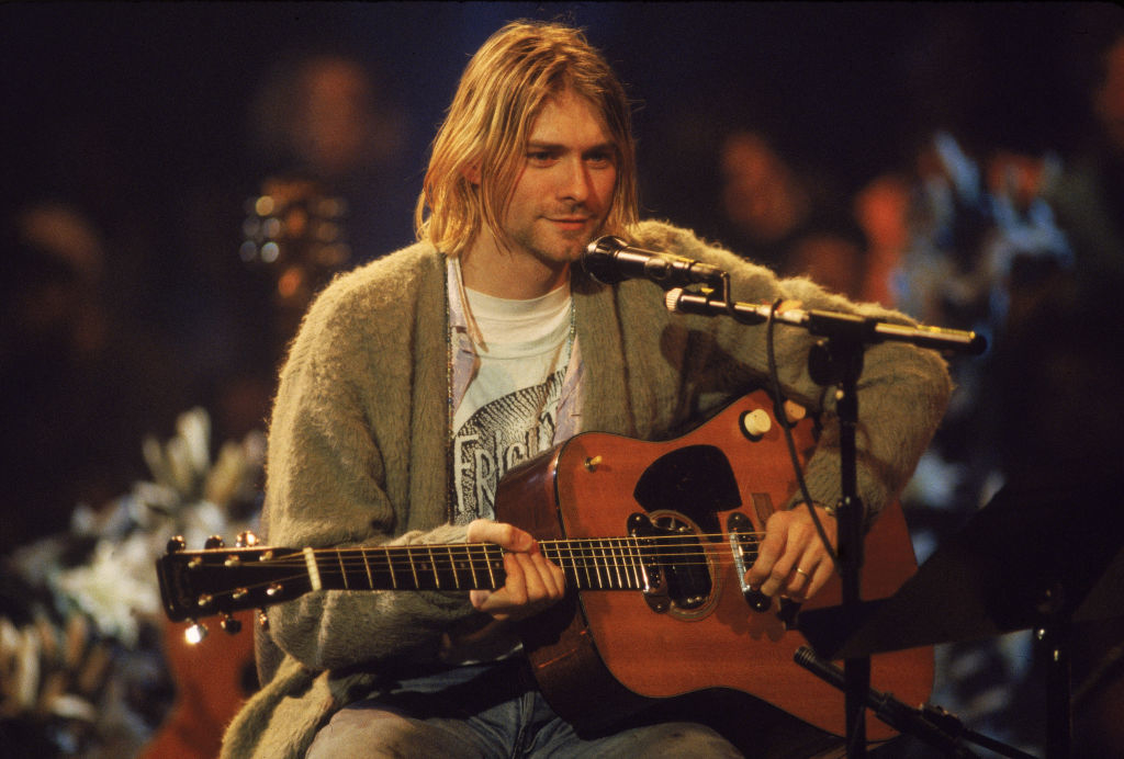 Kurt Cobain playing an acoustic guitar while seated. He is wearing a casual outfit with a cardigan and a t-shirt. A microphone is positioned in front of him