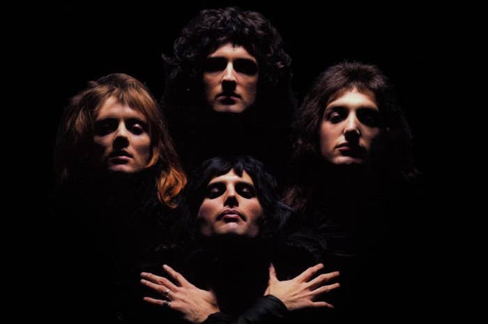Queen band members in a dark, dramatic portrait with their hands and faces illuminated: Freddie Mercury, Brian May, Roger Taylor, and John Deacon