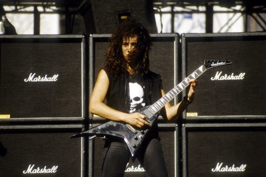 A person with curly hair plays an electric guitar in front of Marshall amplifiers on a stage. They wear a sleeveless shirt with a skull graphic