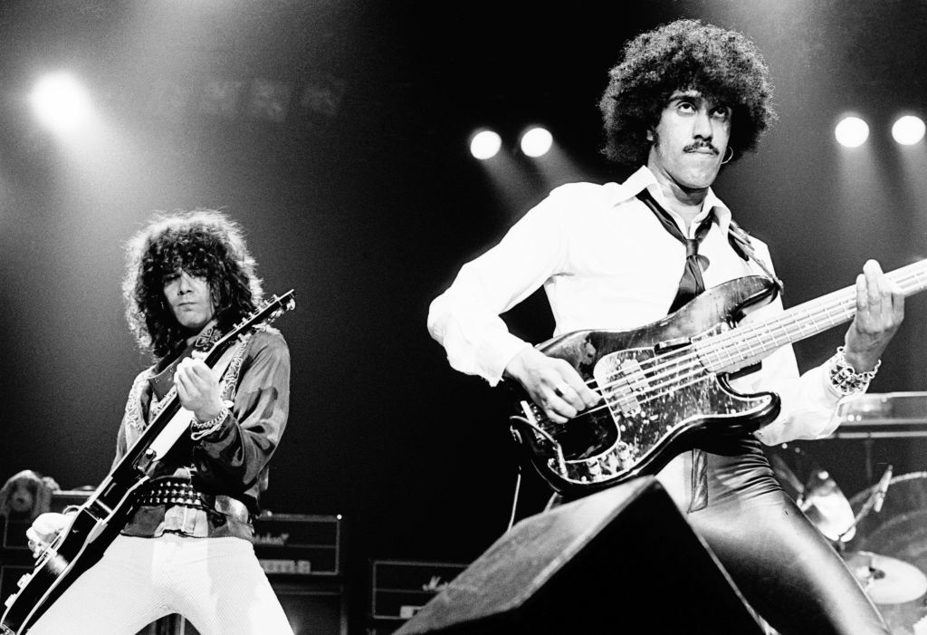 Two musicians perform on stage. The guitarist wears a dark shirt and the bassist in a white shirt and dark pants. Instruments and lights are visible in the background. Names not known