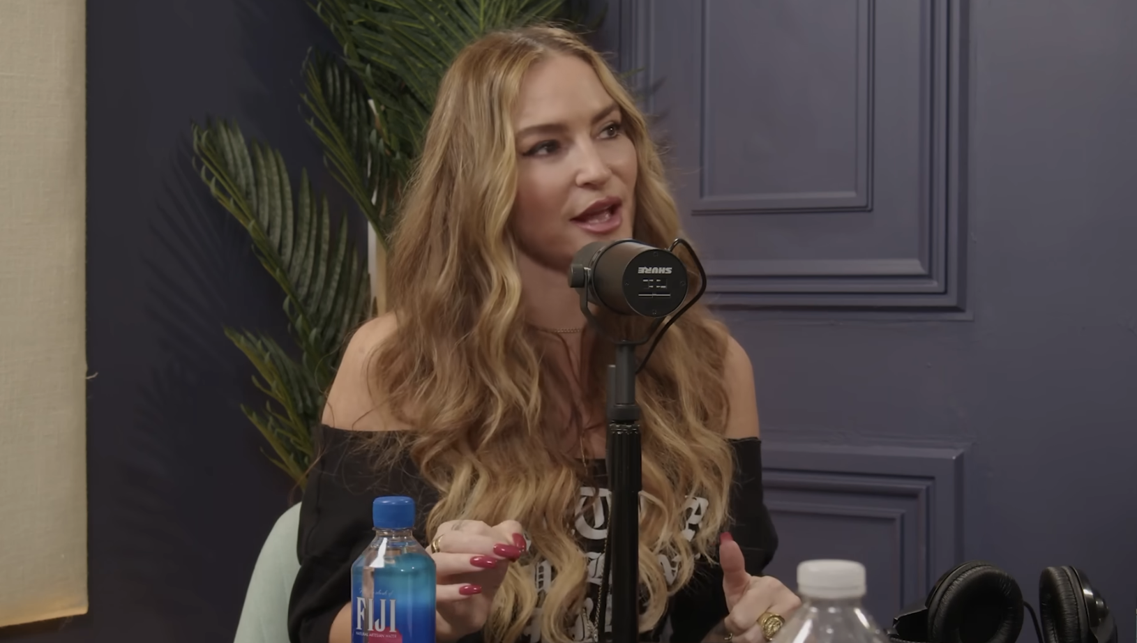 Drea de Matteo with long wavy hair speaking into a microphone in a recording studio, with a bottle of Fiji water in the foreground.