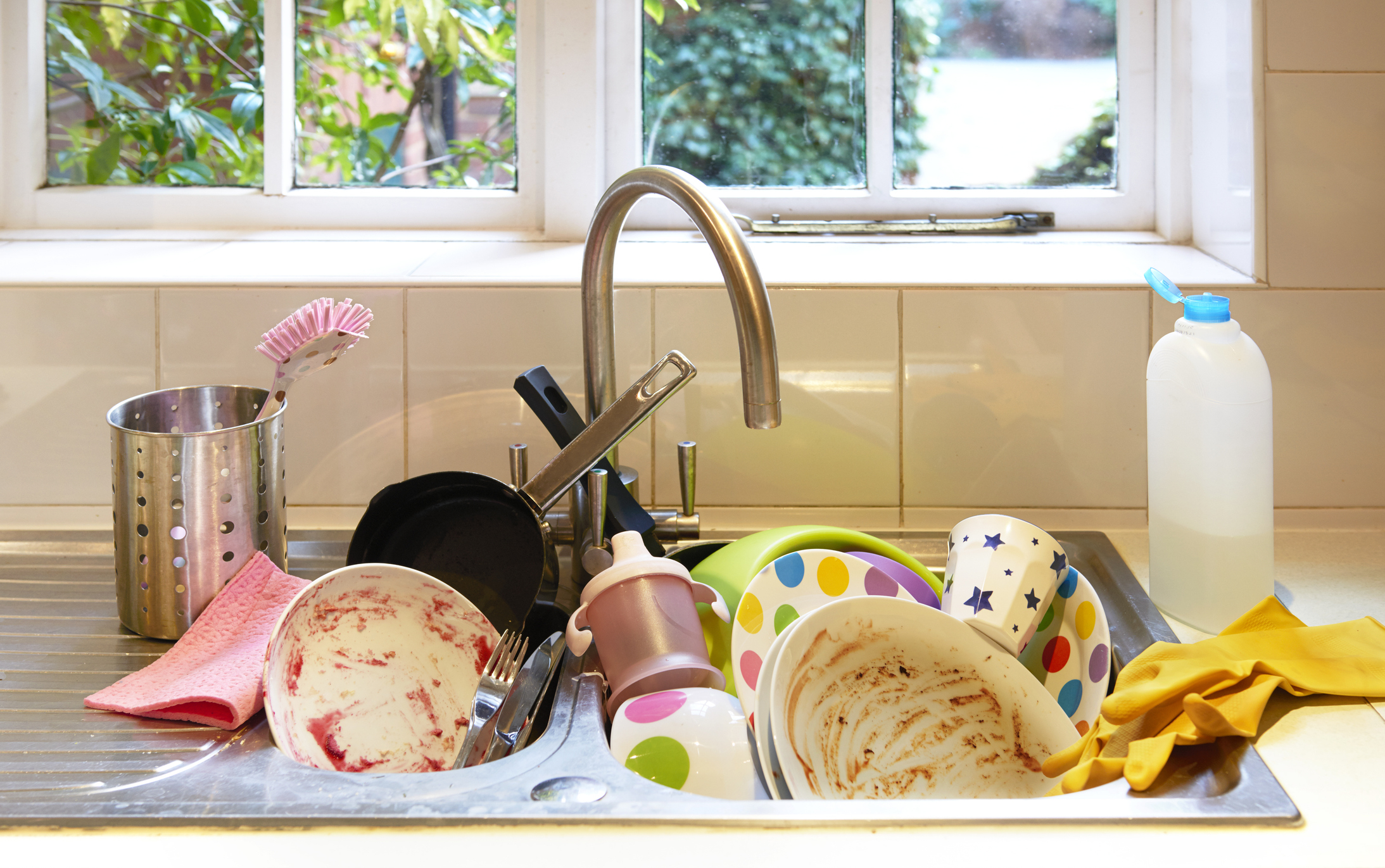 A kitchen sink filled with unwashed dishes, utensils, and a bottle, next to a window with greenery outside