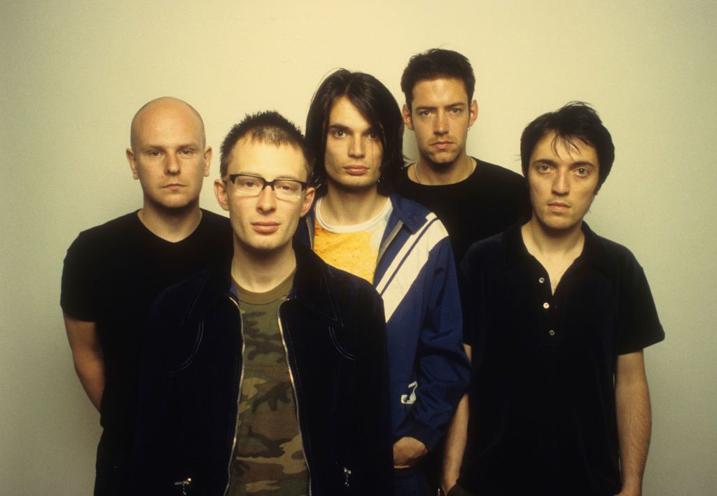 Five men, Thom Yorke, Jonny Greenwood, Colin Greenwood, Ed O'Brien, and Phil Selway, stand together facing the camera. They appear serious