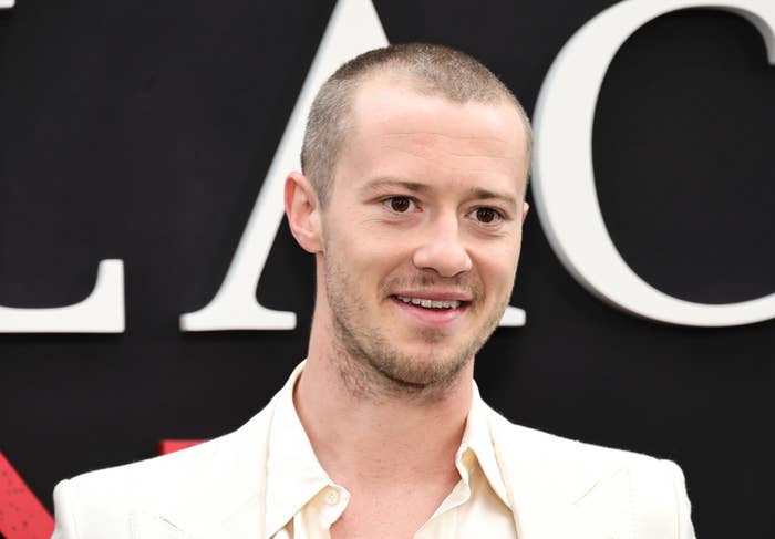 Joseph Quinn is smiling, wearing a light-colored suit jacket and shirt, standing in front of a dark background with large, partially visible letters