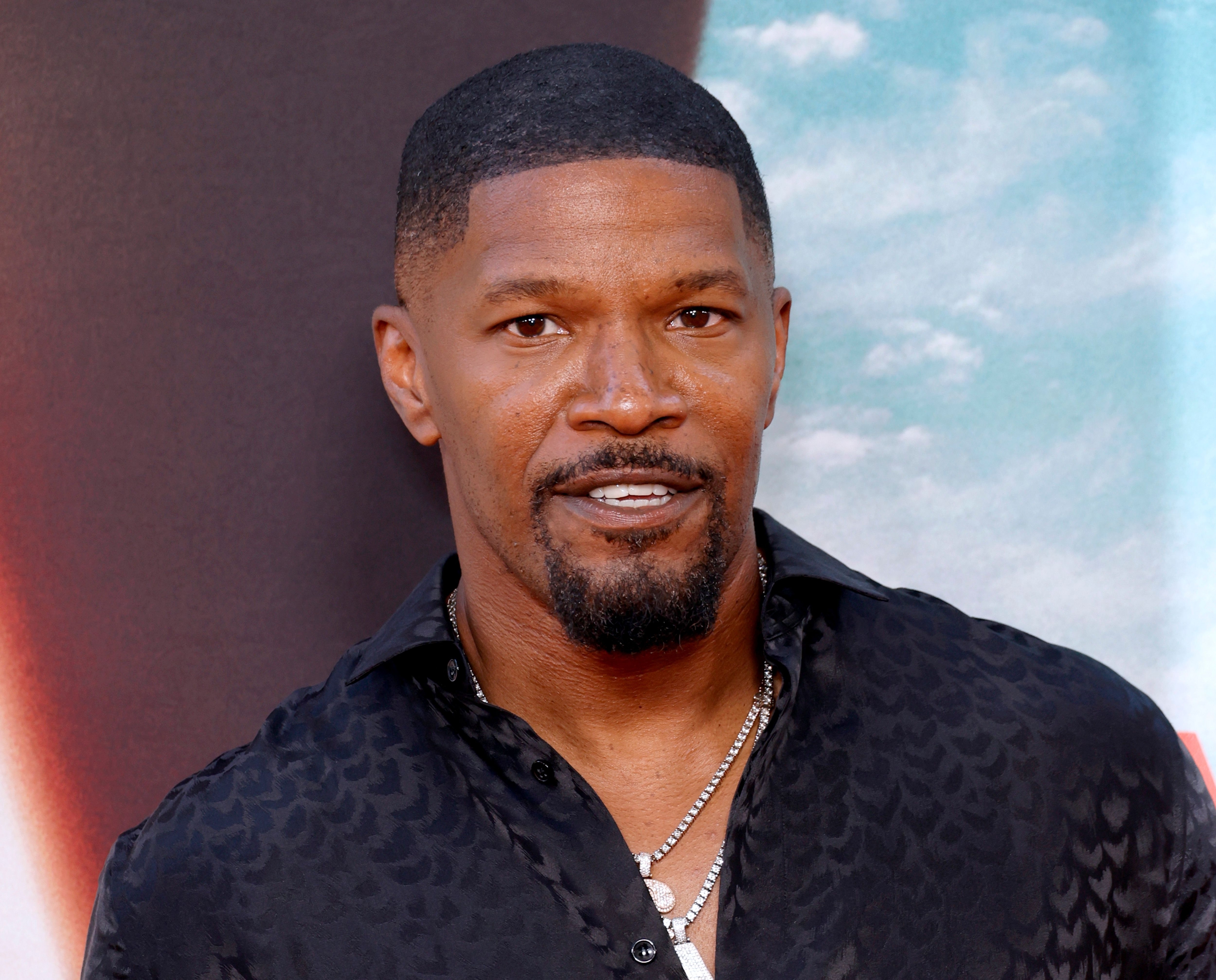 Jamie Foxx on the red carpet, wearing a patterned black shirt and layered silver chains, smiling at the camera