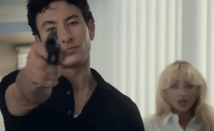 Barry Keoghan points a gun directly at the camera while Sabrina Carpenter looks scared in the background in a scene from the music video for “Please Please Please”