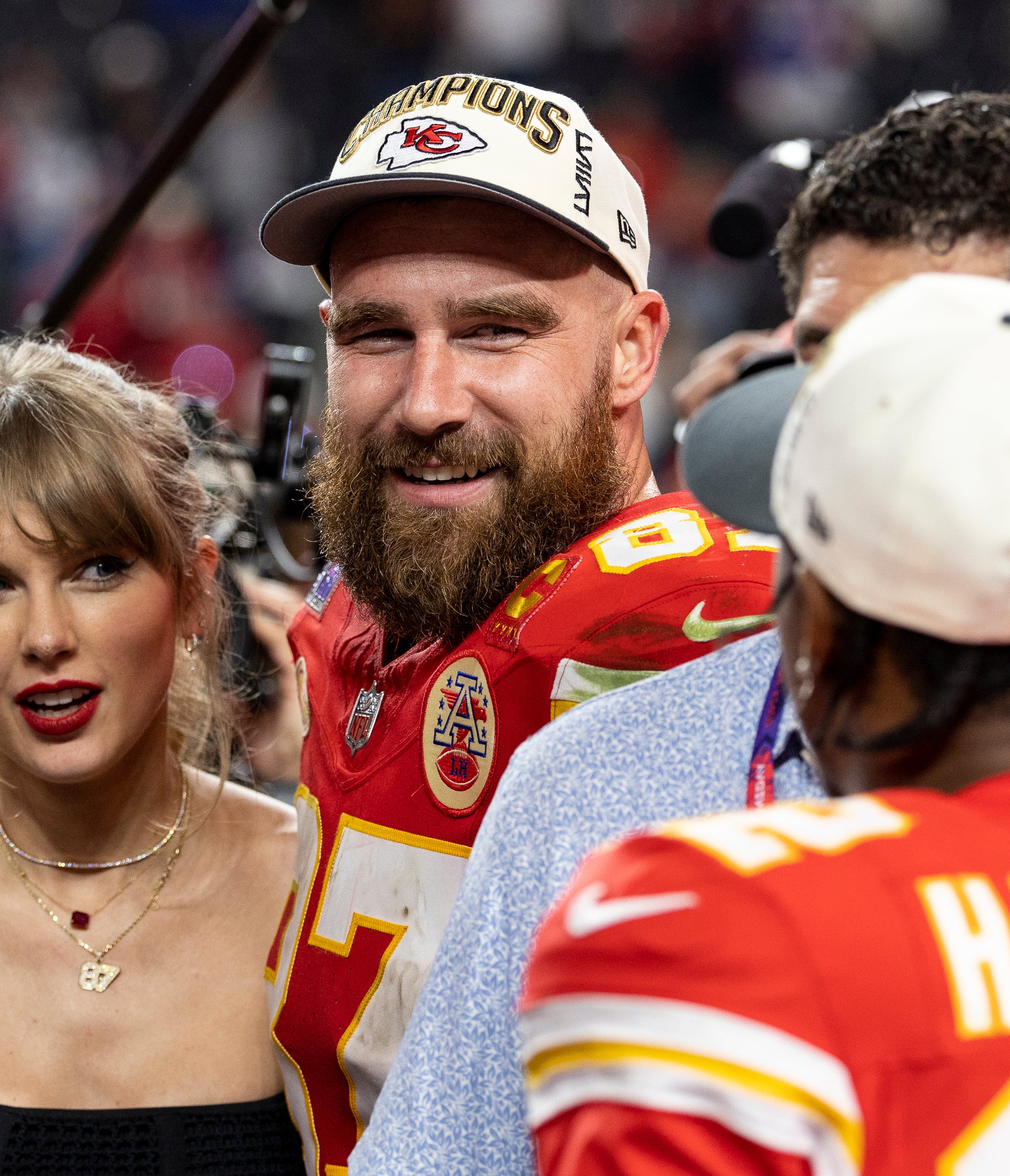 Taylor Swift and Travis Kelce smiling together surrounded by people and media cameras. Travis is in a football uniform and championship cap, standing near Mecole Hardman