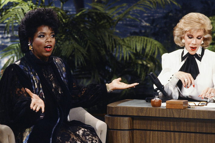 Oprah Winfrey, wearing a patterned dress, gestures while talking to Joan Rivers, who is seated at a desk during a TV interview on a talk show set