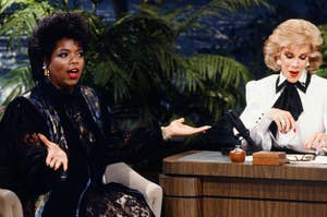 Oprah Winfrey, wearing a patterned dress, gestures while talking to Joan Rivers, who is seated at a desk during a TV interview on a talk show set