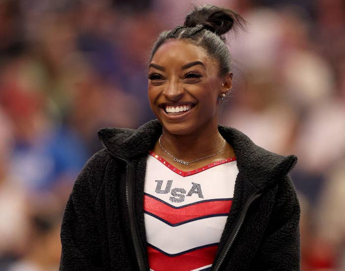 Simone Biles smiles on the sidelines, wearing a leotard with "USA" on the front and a jacket on top
