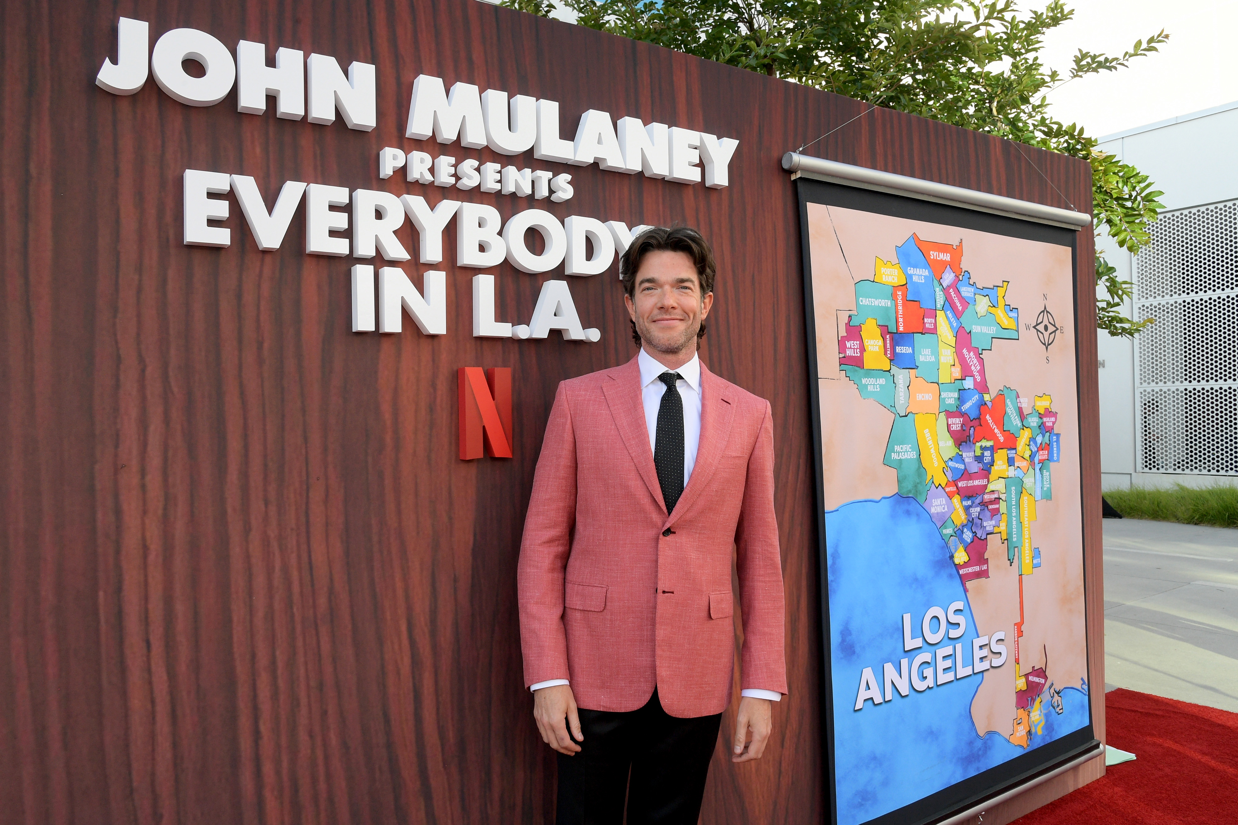 John Mulaney stands in front of a backdrop that reads "John Mulaney presents Everybody in L.A." and a colorful map of Los Angeles. He is wearing a pink suit