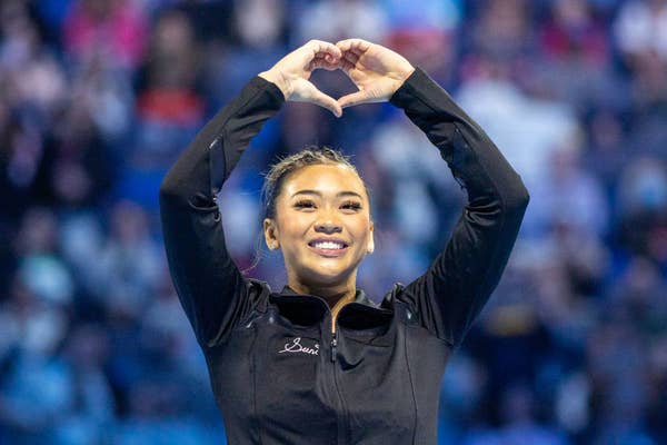 Suni Lee on stage, smiling and making a heart shape with her hands over her head. She is wearing a black athletic jacket