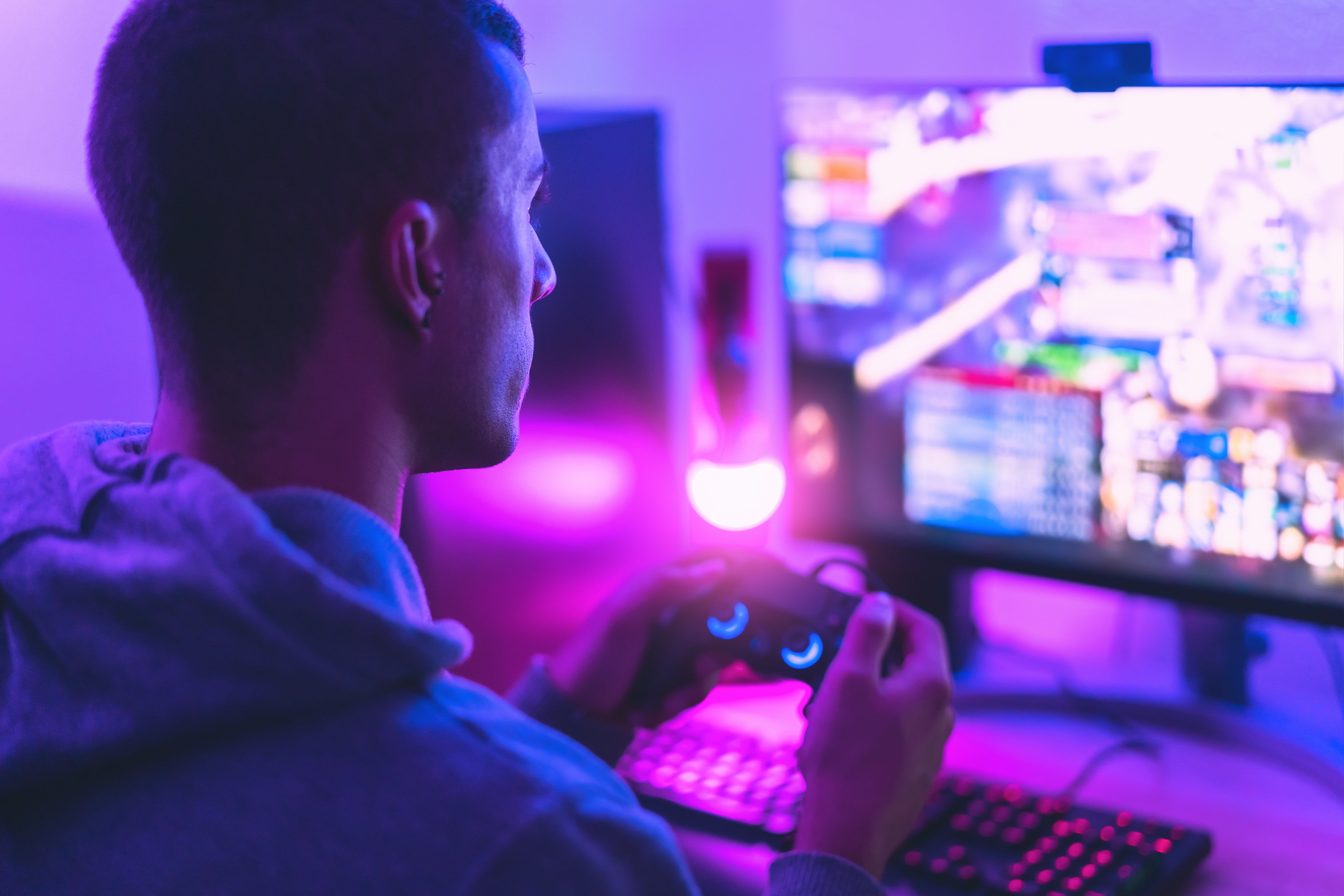 A person is playing video games on a computer setup with a controller in hand, illuminated by ambient purple and blue lighting