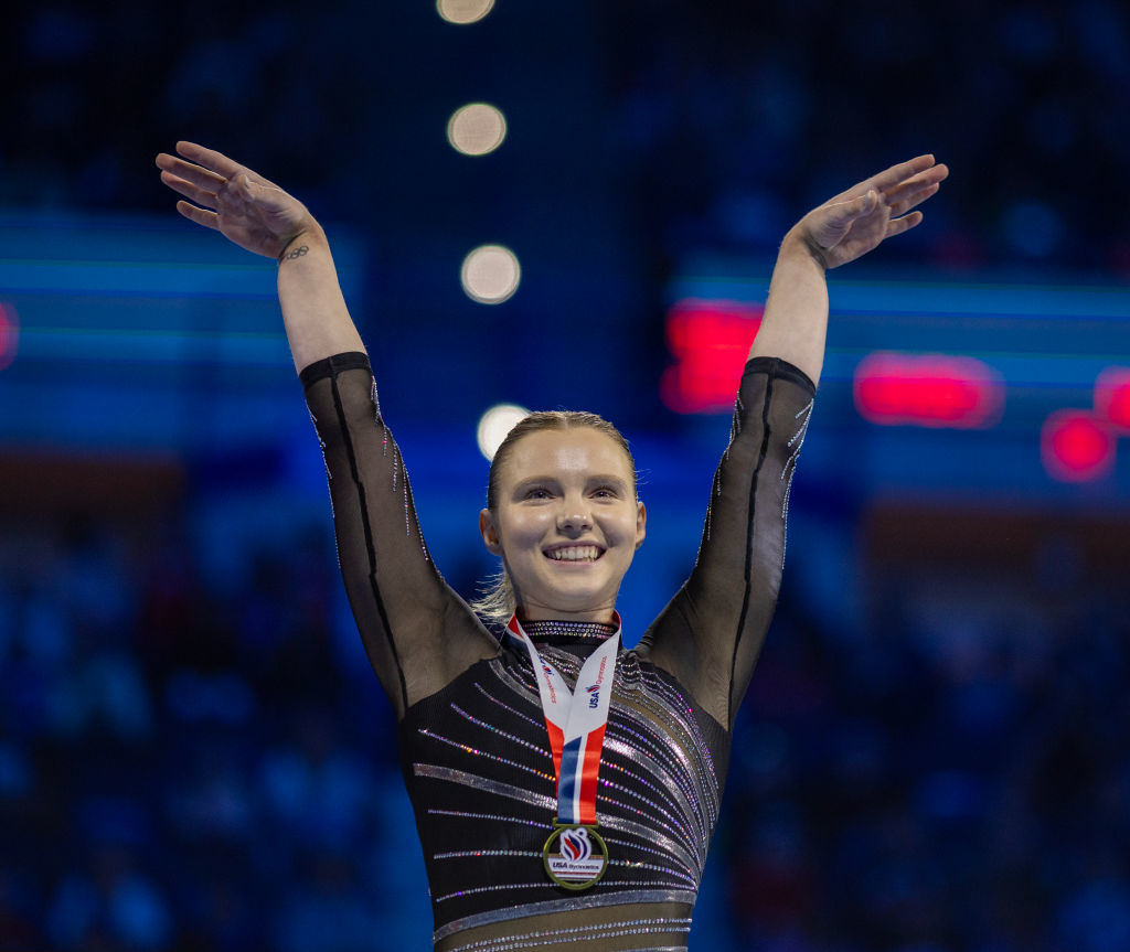 Jade wearing a dark, long-sleeve leotard celebrates with arms her raised, while wearing a medal around her neck in an indoor arena