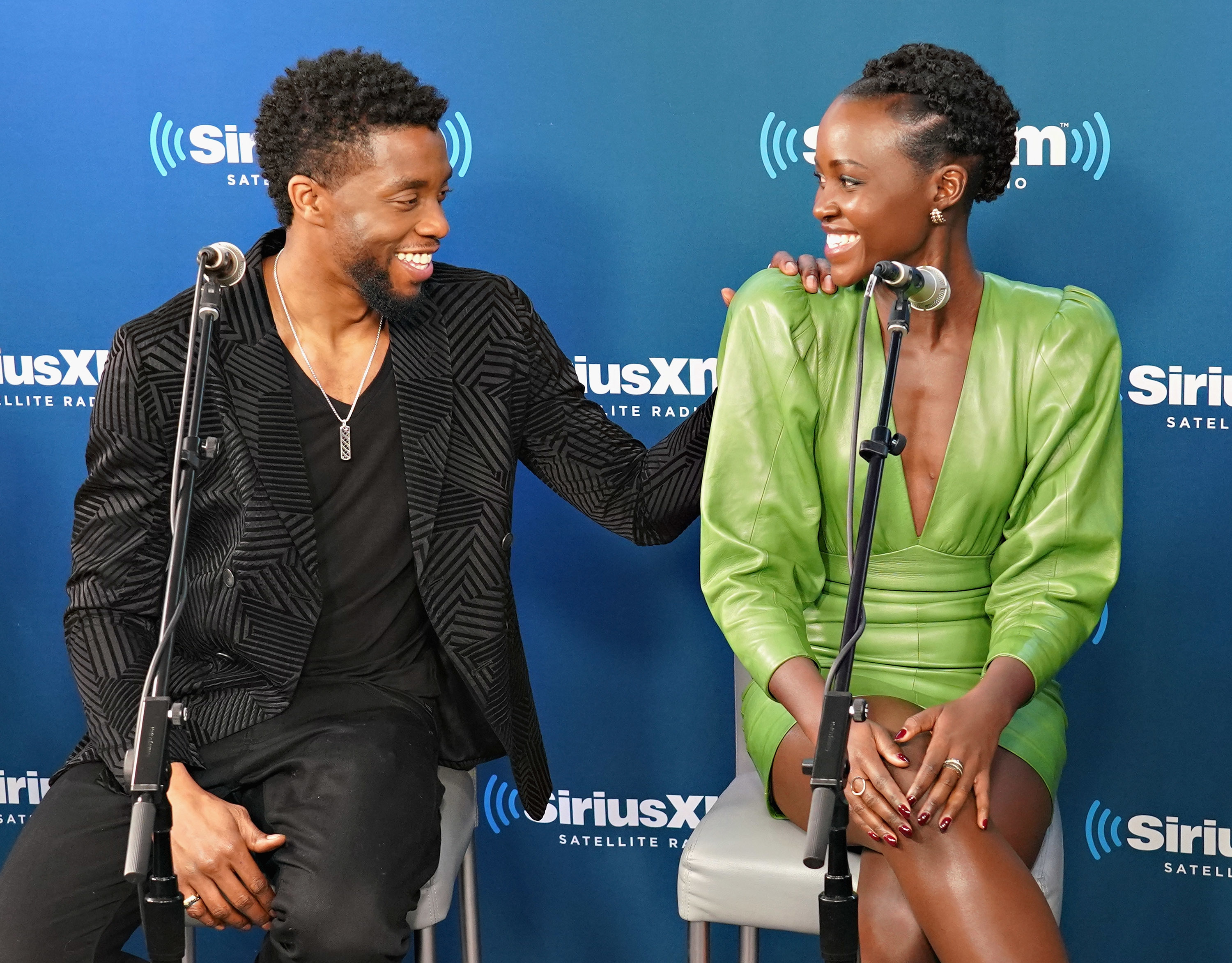 Chadwick Boseman and Lupita Nyong&#x27;o seated at SiriusXM event, Chadwick wearing a blazer, Lupita in a dress, both smiling and looking at each other