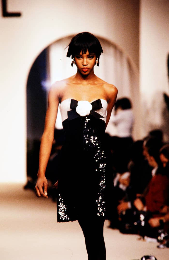 A younger Naomi Campbell on the runway, wearing a strapless dress with a white flower and black bow, adorned with sequins