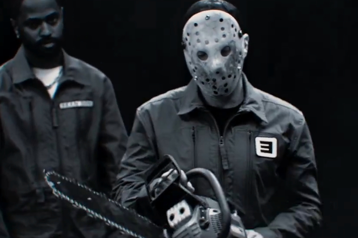 Eminem dressed as Jason Voorhees holding a chainsaw, flanked by Big Sean and another individual in jumpsuits, in a horror-themed music video scene