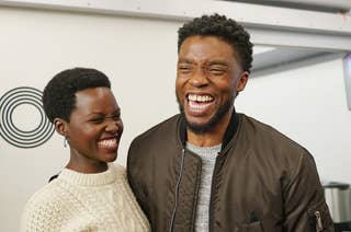 Lupita Nyong'o in a sweater and Chadwick Boseman in a jacket laughing together indoors