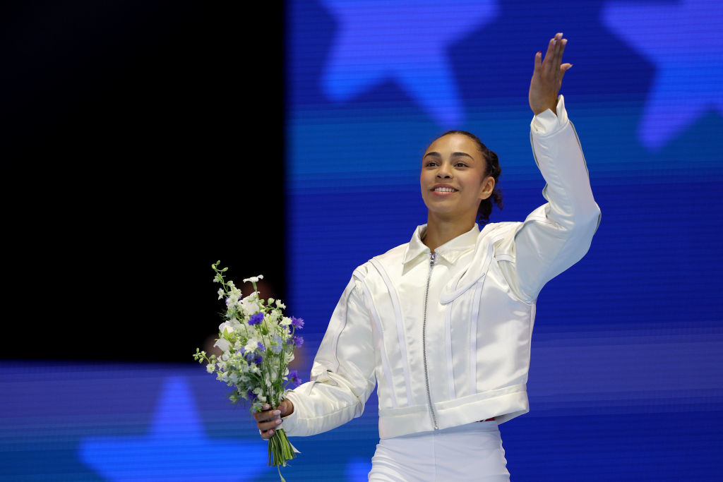 Hezly Rivera, dressed in a white jacket, waves while holding a bouquet of flowers