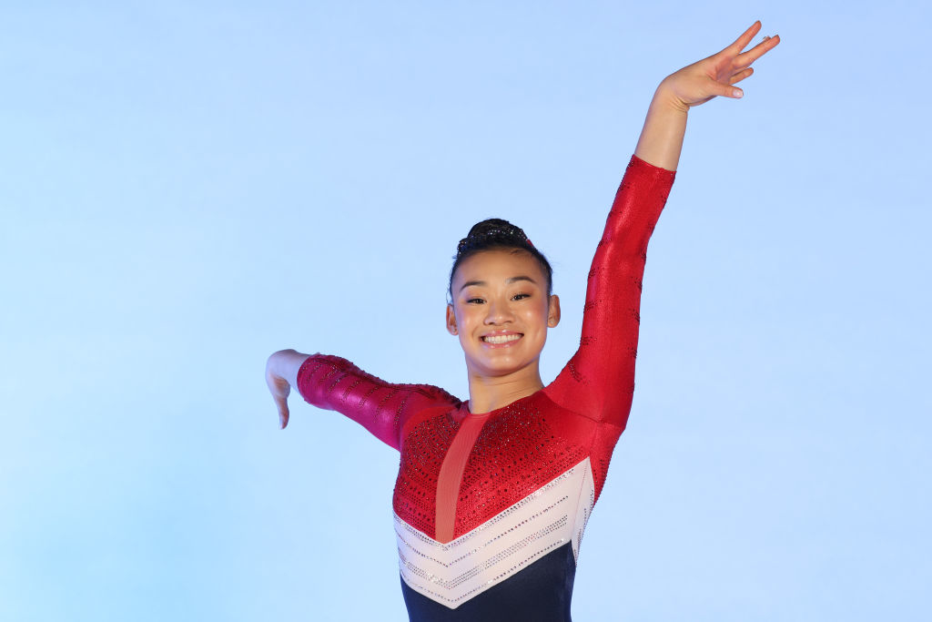 Leanne Wong poses in a red and black gymnastics leotard with one arm raised above her head and the other extended to the side, smiling widely