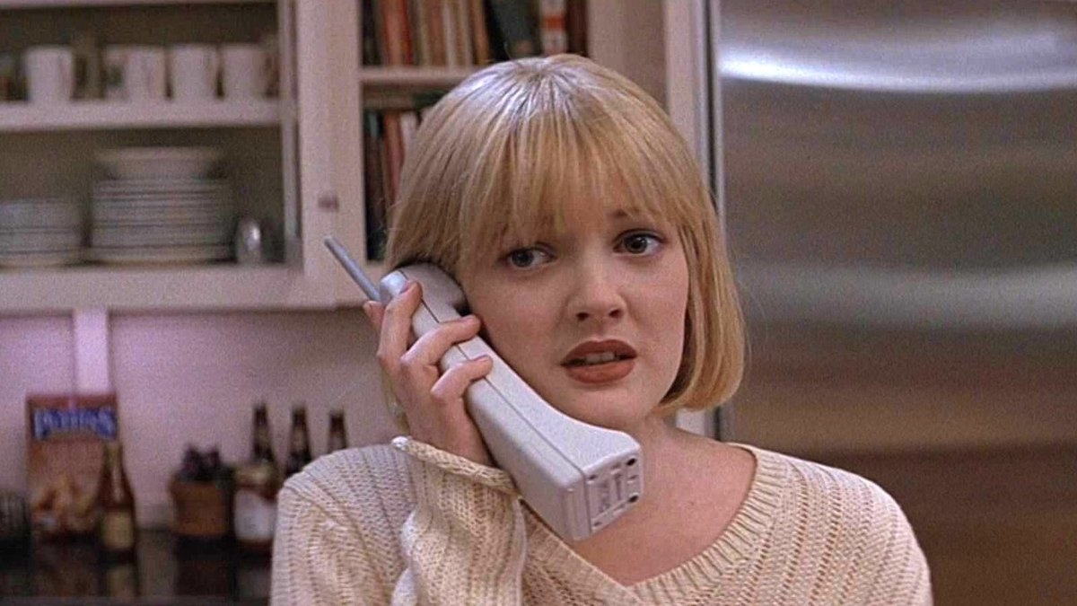 Drew Barrymore, in a cream sweater, holds a large cordless phone to her ear in a kitchen scene. She looks worried or scared