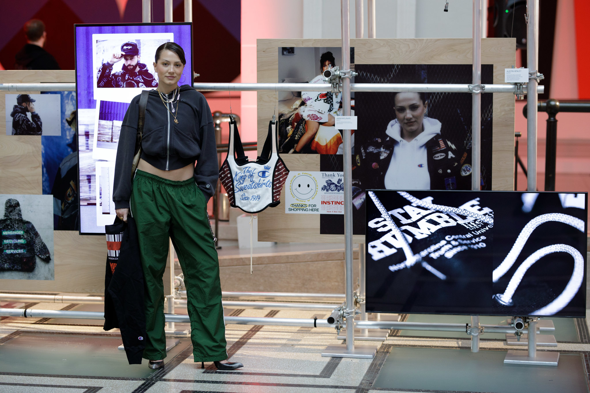 Bella Hadid poses at a fashion exhibition, wearing a cropped hoodie and green cargo pants. Various fashion displays and images surround her in the background