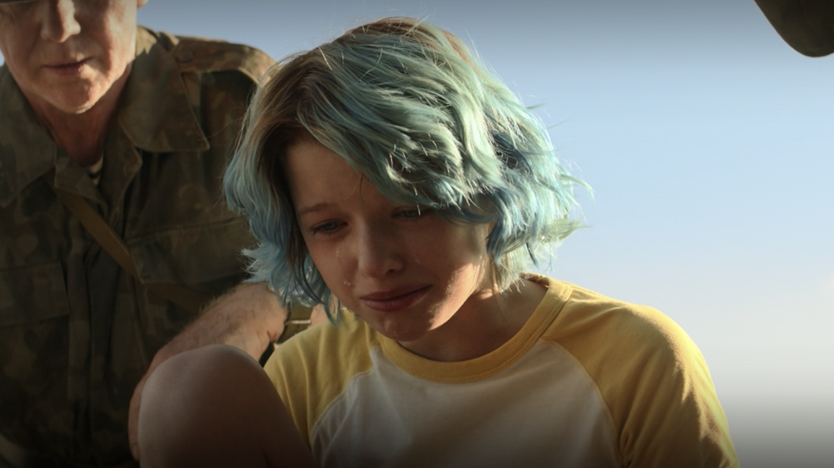 A young person with blue hair, wearing a yellow and white shirt, looks sad and teary-eyed, with an older person in military attire in the background
