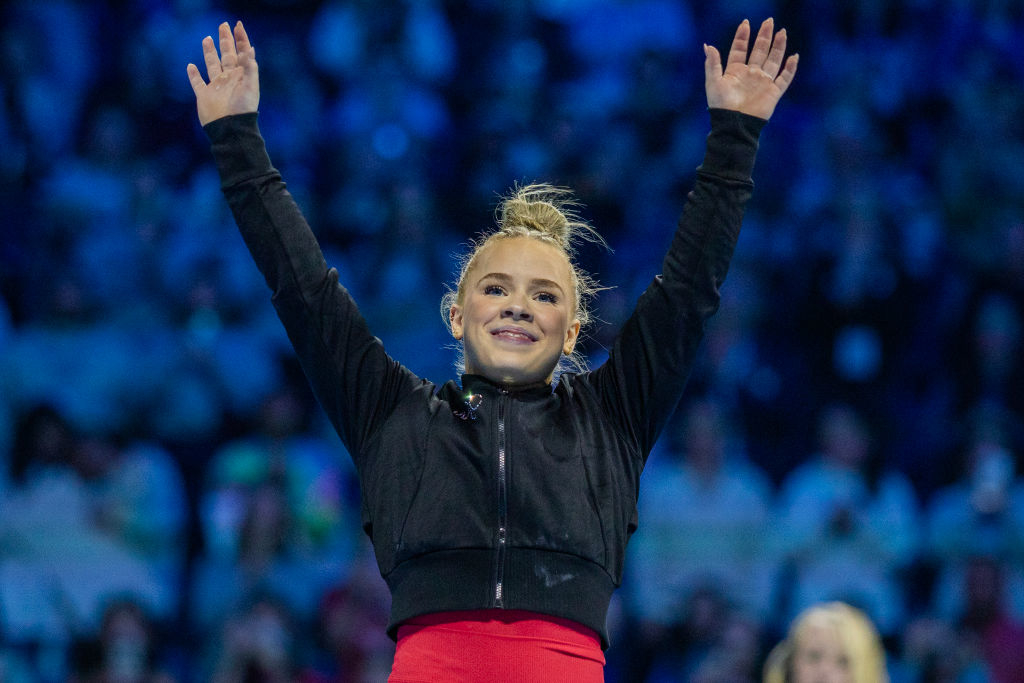 Joscelyn stands with arms raised in triumph at a sports event, wearing a black jacket and red pants