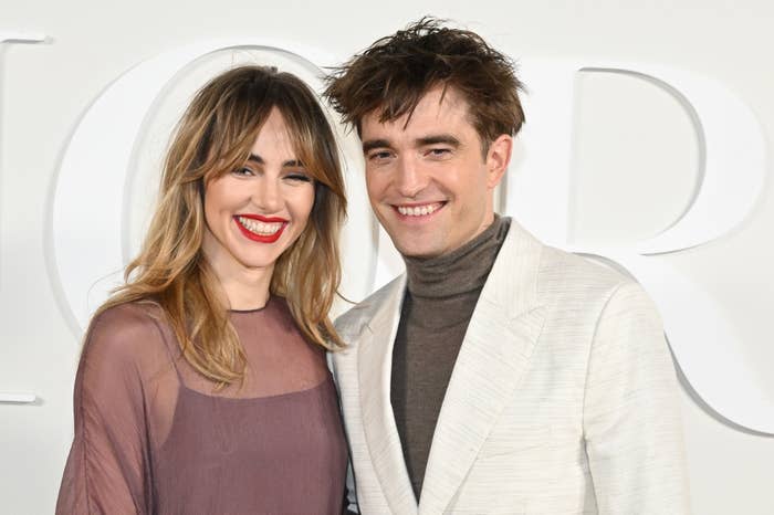 Suki Waterhouse and Robert Pattinson pose together. Suki wears a sheer top, and Robert wears a light-colored suit with a turtleneck