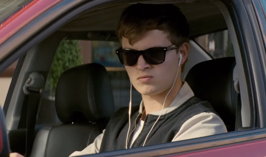 Ansel Elgort is driving a car, wearing sunglasses and earphones, in a scene from the film "Baby Driver."
