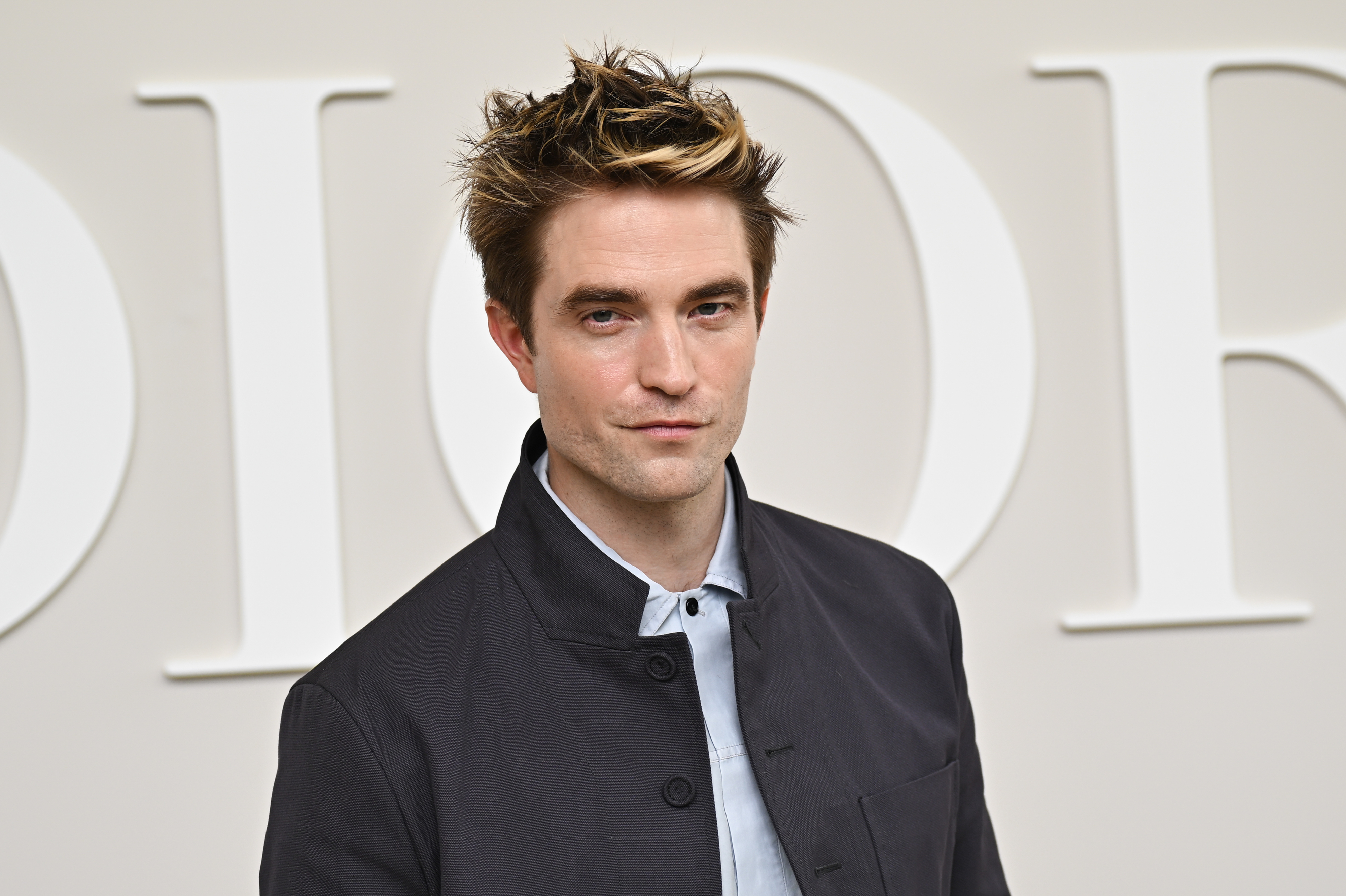 Robert Pattinson poses in a button-up shirt and jacket at an event with large "Dior" signage in the background