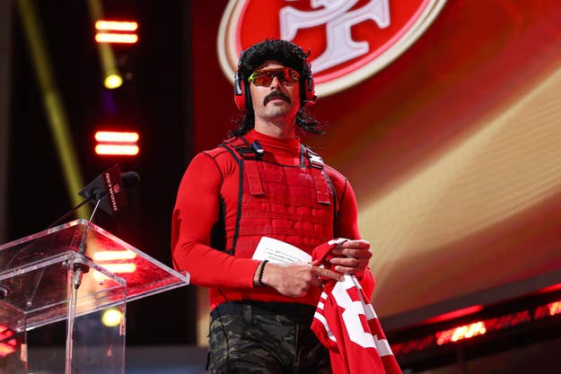 Dr Disrespect wears a red shirt, tactical vest, and sunglasses while holding a football jersey and standing at a clear podium, under a lighted stage backdrop