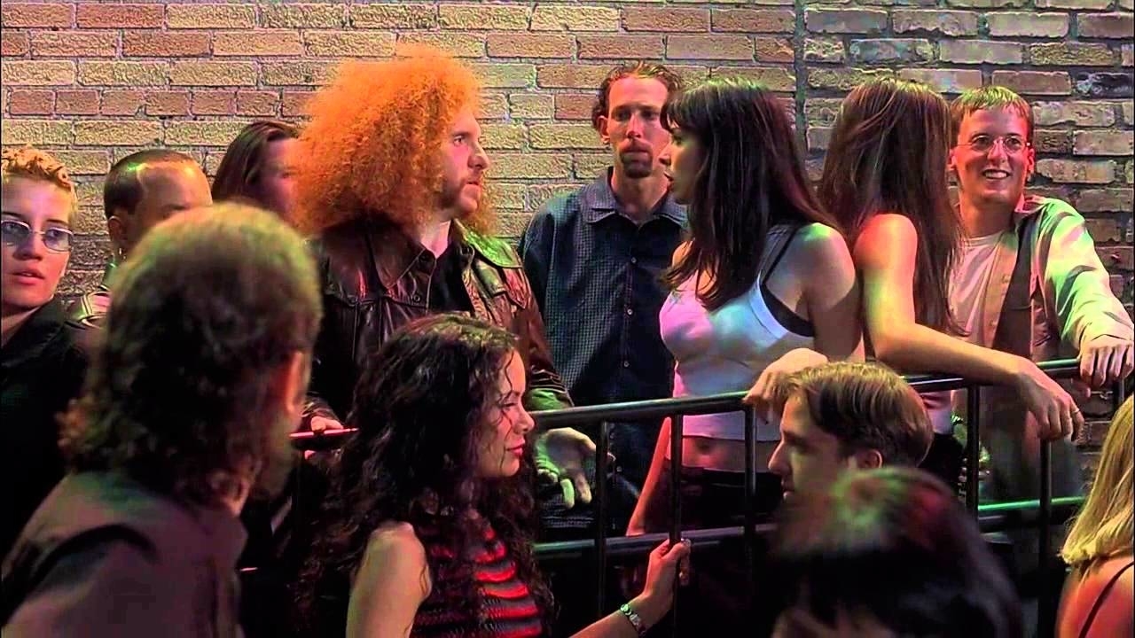 A group of people in casual clothes converse and mingle at a crowded event against a brick wall. They seem to be enjoying their time