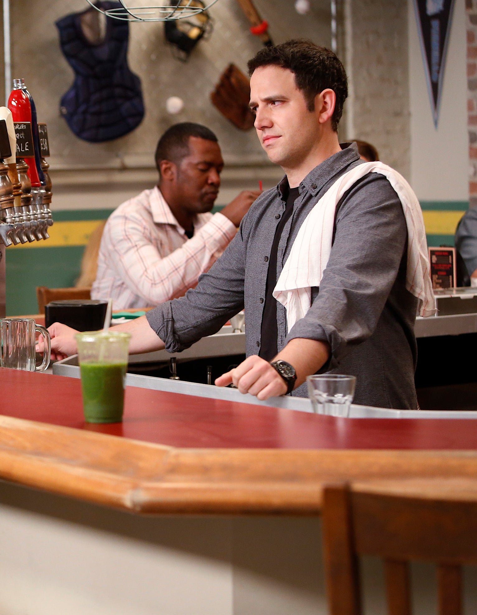 Greg behind a diner counter, with another patron seated further back. Various sports items are displayed on the wall