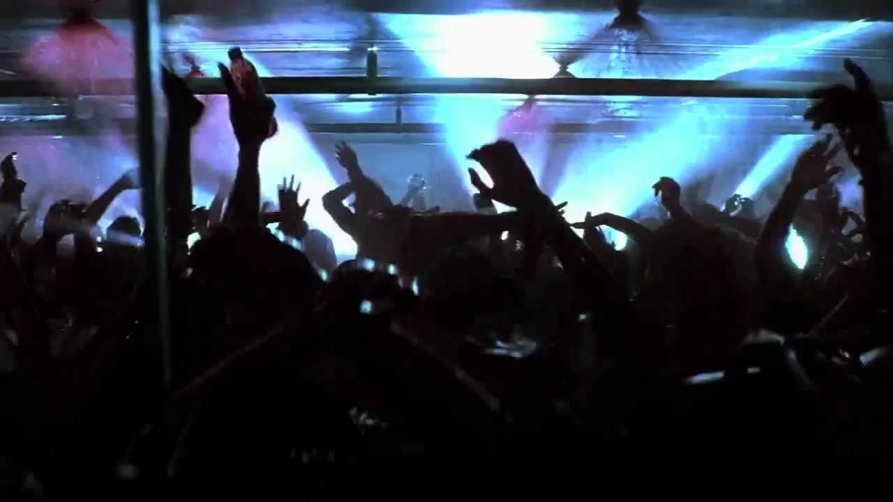 A crowded, energetic dance floor with people raising their hands and dancing under bright, flashing lights in a nightclub